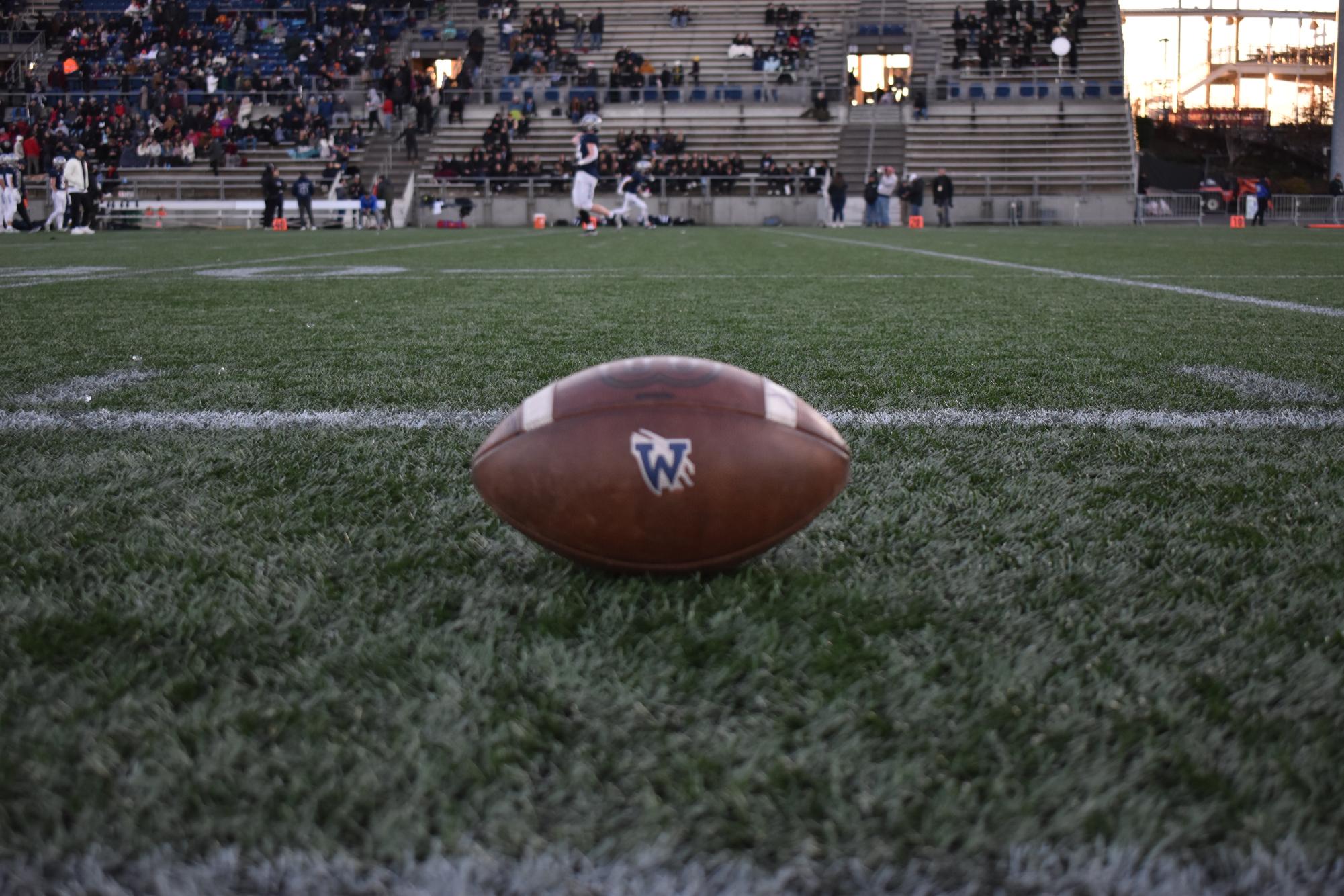 A Wilsonville football lays on the sideline as the team warms up in the background. The State Final matchup started at 4:30 p.m. with a beautiful sunset as the backdrop for the game.