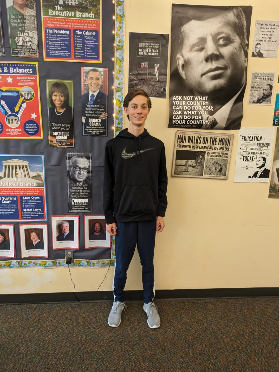 Wilsonville student Malachi Tadema poses in front of posters in Mr. Navarro's classroom. The wall shows a display of influential people in history, including John F. Kennedy, Michelle Obama, and more.