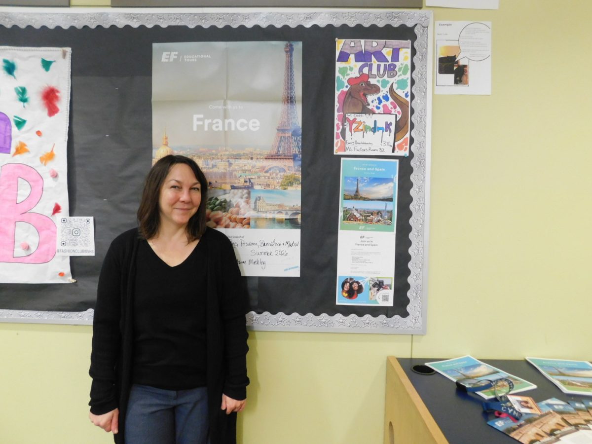 Ms. Moehling stands in front of the French trip poster that's on its way to being planned. She's smiling happily, excited to get to show her students around France!