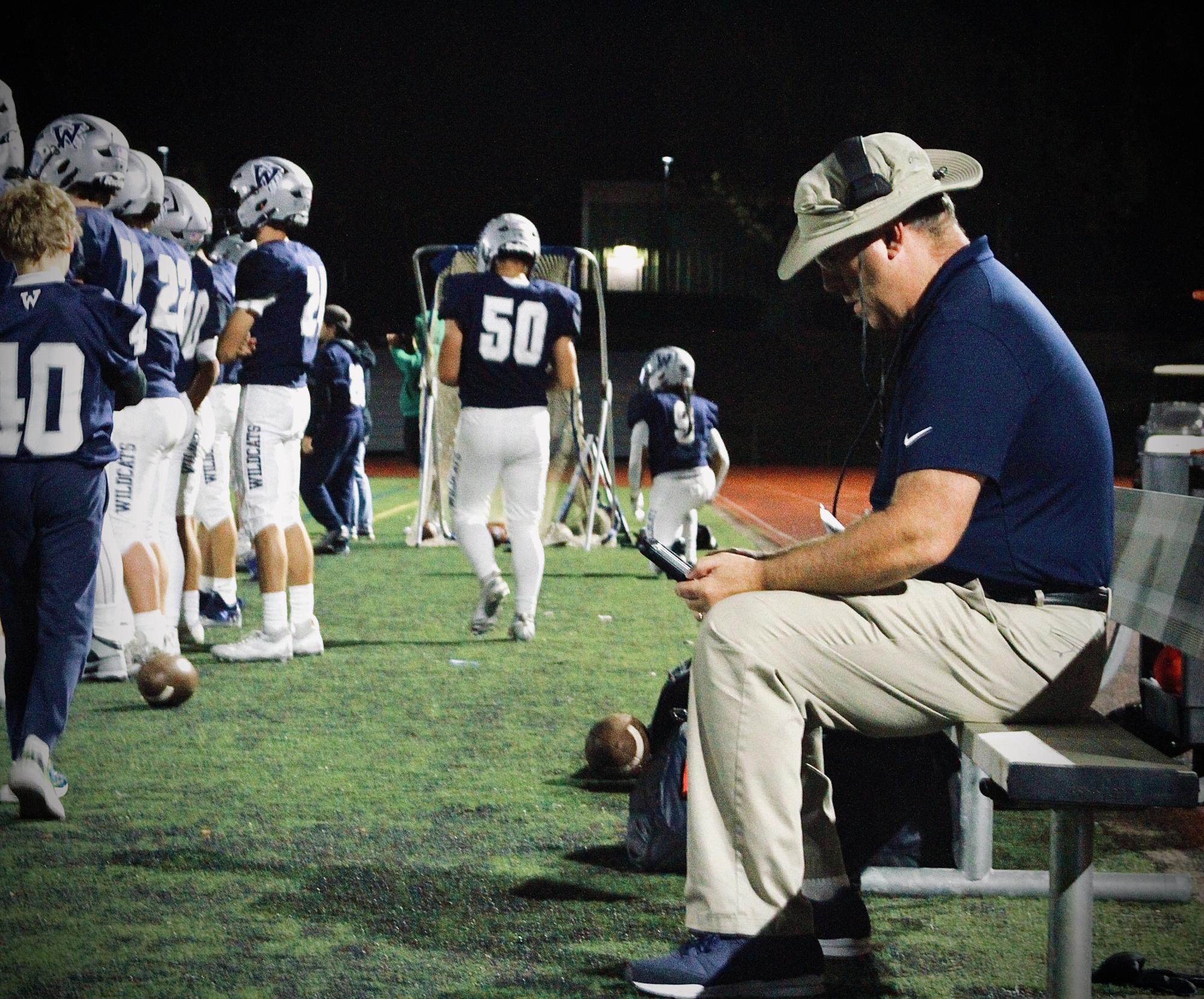 Coach Guenther takes a seat on the player bench behind his sideline while glancing over plays. Headset gear allows for smooth communication during games between players and coaches. 