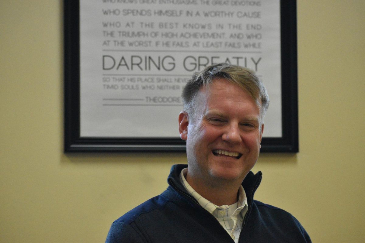 Jeff Deeder seated in his classroom, grading assignments in front of a quote by President Roosevelt. As an economics teacher, Deeder has found joy in creating a classroom environment for his students to thrive.