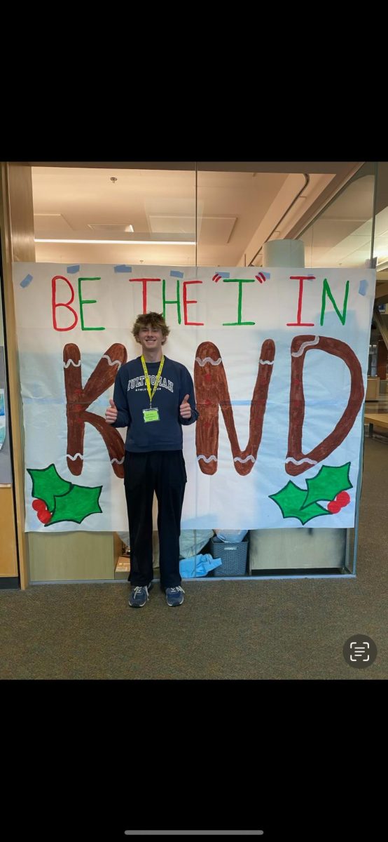 Senior Nick Waite poses in front of a festive sign about kindness. It's important to be kind to everyone.