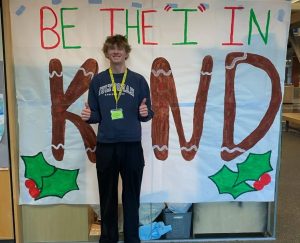 Senior Nick Waite poses in front of a festive sign about kindness. It's important to be kind to everyone.