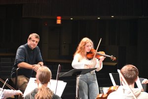 Wilsonville High School band and orchestra director Chad Davies smiles in appreciation as chamber orchestra concertmaster Ellie McClelland leads the orchestra in the string-tuning process. Student leaders are a cornerstone of Wilsonville’s music program. Photo taken by the author.