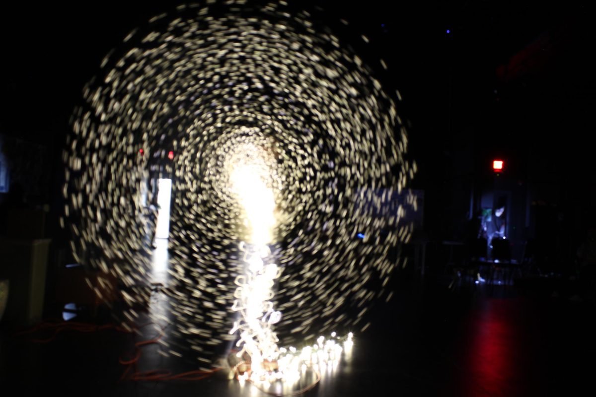 Andrea Ormsby and her class take photos of the display of lights set up in a dark room. She lights burst down through the middle and make a swirl like figure.